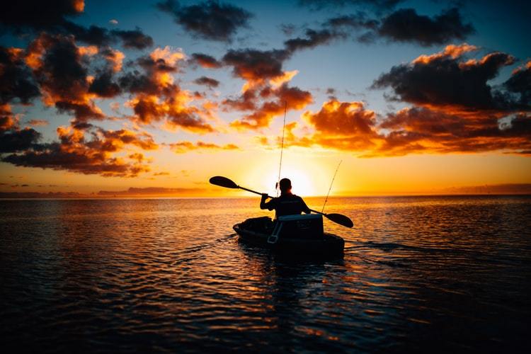 silhouette of man with fishing gear in kayak on water at sunrise