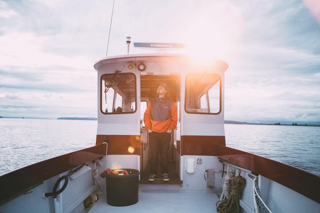 man wearing orange jumper and dark pants standing in cabin of fishing boat on water